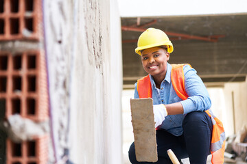 Full length portrait of happy young female labor with trowel crouching on plank by wall at construction site