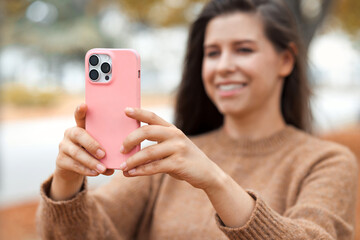 Young happy woman take a pictures of autumn nature in the park with her new smartphone. Pink silicone protective case on mobile phone. 
