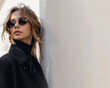 © Qstock - Stylish Young Woman Wearing Sunglasses and Black Coat Leaning Against Wall - Fashion Portrait