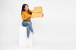 © comzeal - Happy Young Asian woman sitting on white box and holding package parcel box isolated on white background, Delivery courier and shipping service concept