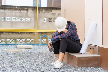 Asian female construction worker sits on steps outside house, wearing safety helmet, holding tablet and resting head on arms, blueprint and coffee cup beside her, taking a break from inspection.