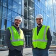 © Tadeusz - Two male construction workers in safety gear smiling at a modern building site.