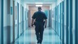 © Bonsales - Caucasian male police officer in uniform with gun on his belt walking down a brightly lit hospital hallway with back turned