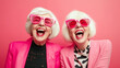 © Poj - Two elderly with colorful dresses and fancy sunglasses laughing with happiness against pale red background.