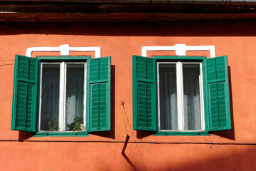 Two green wooden shutters partially open on a bright orange wall. The windows behind the shutters have white frames and are adorned with sheer curtains.