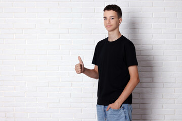 Wall Mural - Teenage boy wearing black t-shirt and showing thumbs up near white brick wall, space for text