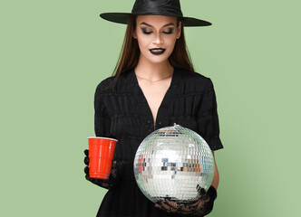 Wall Mural - Young woman dressed for Halloween as witch with drink and disco ball on green background