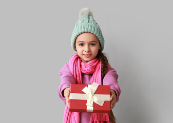 Cute little girl in warm hat and scarf with Christmas gift on grey background