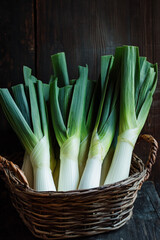 Wall Mural - A basket of organic leeks, with their long green leaves and white stalks, placed against a dark wood background, showcasing their fresh, crisp texture.