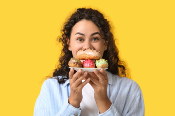 Wall Mural - African-American woman with tasty eclairs on yellow background