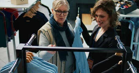 Wall Mural - Retail worker suggesting fashion items to elderly customer in clothing store, offering various clothing choices to extend wardrobe. Regular client discussing about style with mall employee. Camera B.