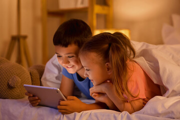 Cute happy little kids with tablet playing video games in evening at home