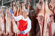 © JackF - Smiling young female butcher shop worker checking fresh raw dressed lamb carcasses hanging on hook frame in cold storage room