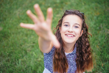 High angle view of a beautiful little girl smiling happily, reaching her hand towards the camera as if she is going to take it. Cute little girl sitting in a park on green grass