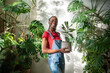 © DimaBerlin - Smiling African American woman holds potted ficus, surrounded by lush flowers and plants in home garden. Black female florist caring of plants, Indoor greenery, hobby, nature lover, mindful living