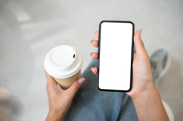 Wall Mural - Top view of a woman in jeans sitting in a cafe, holding a coffee cup while using her smartphone.