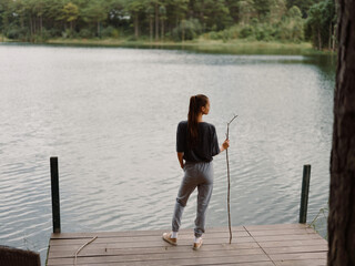 Thoughtful woman standing by the lake, holding a stick, contemplating nature and solitude Peaceful outdoor scene with trees reflecting on water