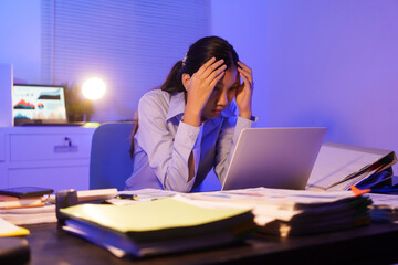 A young woman works overtime at night at her desk in office, using a laptop reviewing documents. is stressed, tired,experiencing headaches,body aches from sitting too long,leading to office syndrome