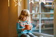© Irina Schmidt - Little preschool girl holding cup with hot chocolate with marhsmallows. Happy child drinking sweet cocoa by window with Christmas lights in winter. Cozy family celebration of xmas.