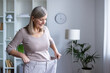 © Liubomir - Happy senior woman examines her weight loss progress by wearing oversized pants. She stands in modern room, proud of healthy lifestyle and personal achievement in fitness journey.