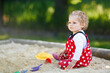 © Irina Schmidt - Cute toddler girl playing in sand on outdoor playground. Beautiful baby in red gum trousers having fun on sunny warm summer day. Child with colorful sand toys. Healthy active baby outdoors plays games
