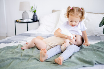 sisters are feeding baby milk from a bottle on the bed at home, the older sister is holding the baby at home on the bed, the concept of baby food