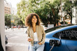 © Jordi Salas - A woman is smiling as she stands next to her electric car at a charging station in a city environment. The scene captures modern transportation and eco-friendly living.