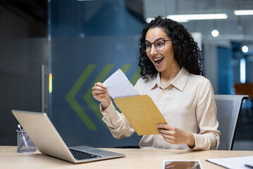 Wall Mural - Businesswoman at desk receives good news in envelope, showing excitement and joy. Seated with laptop and phone in office setting.