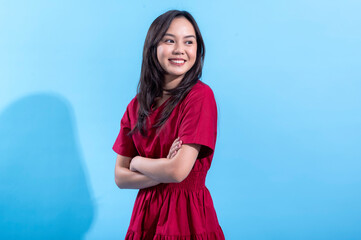 A young Asian woman smiles confidently, arms crossed, wearing a bright red dress against a light blue background. Her casual yet stylish look, combined with the vibrant colors, creates a positive