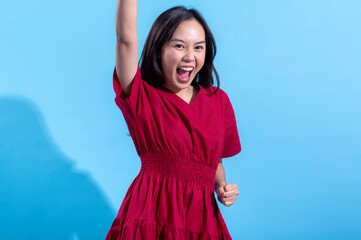 An excited Asian woman in a red dress raises both fists in the air while cheering enthusiastically. She stands against a light blue background, displaying an expression of joy and triumph