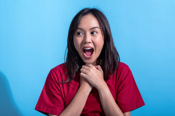 Wall Mural - An Asian woman in a red dress stands against a light blue background with her mouth wide open, expressing shock or surprise. Her hands are clasped near her neck, emphasizing her astonishment