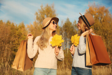 Wall Mural - Happy two women friends with autumn yellow leaves, shopping bags, stylish girlfriends smile in park