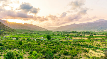 sunset valley landscape with green trees and gardens among beautiful mountains with colorful cloudy sky on background