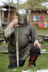 Medieval warrior dressed in chainmail and helmet kneeling on grass holding large sword in an outdoor setting with historical buildings and shields in background