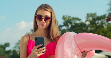 Summer girl using smartphone and sunbathing at swimming pool. Female model relaxing outdoors at sunny day.
