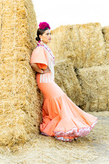 A beautiful woman in her 20s poses in a salmon-colored gypsy or flamenco dress outdoors looking to the side next to a block of straw. Concept of women dressed as flamenco dancers.