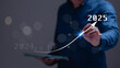 © Mdisk - A man is pointing a graph on a tablet with a blue shirt. The graph shows a line that is rising to the right, with the number 2025 at the top. Concept of progress and growth