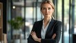 © Nathan - Confident Caucasian businesswoman in a black suit stands with arms crossed in a modern office setting.