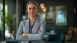 © Nathan - Confident woman with blonde hair in a business suit, standing with arms crossed in a modern office setting.