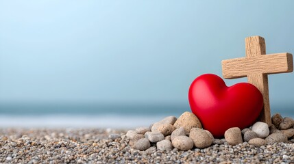 A wooden Christian cross and a red heart resting side by side on a gravel beach with the calming ocean and sky in the background