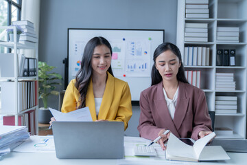 Two young businesswomen are working together in a modern office, analyzing financial documents and using a laptop to input data