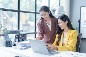 Two asian businesswomen collaborating in a bright office, using a laptop to discuss a project and analyze data and charts, smiling and confident as they work together