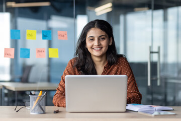 Wall Mural - Young woman with laptop in office setting, smiling confidently, showcasing productive and positive workspace. Surrounded by colorful sticky notes representing ideas and planning.