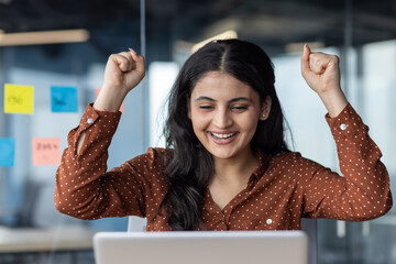 Joyful woman expressing excitement with raised fists, working on laptop in office. Celebrating success or achievement with happiness and enthusiasm