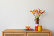 © FollowTheFlow - A striking floral centerpiece of orange calla lilies in a pink and orange ceramic vase, styled with a yellow decorative object and books on a wooden cabinet.