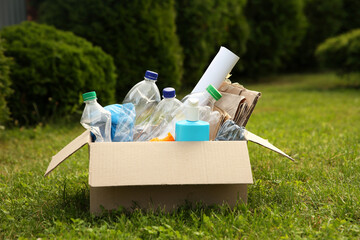 Sticker - Recycling. Different garbage in cardboard box on green grass outdoors