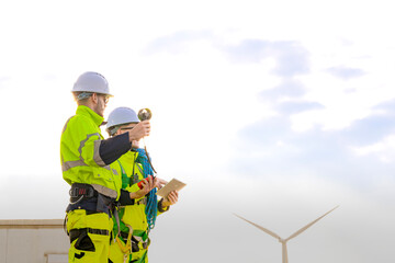 Two engineers wearing high visibility safety jackets and helmets measure wind speed with an anemometer and analyze data on a tablet at a renewable energy site. The cloudy sky adds depth to the scene.