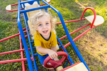 Young boy with curly blonde hair is smiling on a playground swing set shaped like an airplane. Selective focus