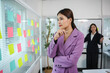 © amnaj - Businesswoman is thinking about solutions for her business, looking at colorful sticky notes stuck on a glass wall