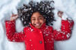 © MarGa - African american girl lying in the snow, laughing joyfully as snowflakes rest on her curly hair and red coat. Concept for winter joy, childhood happiness, and holiday fun